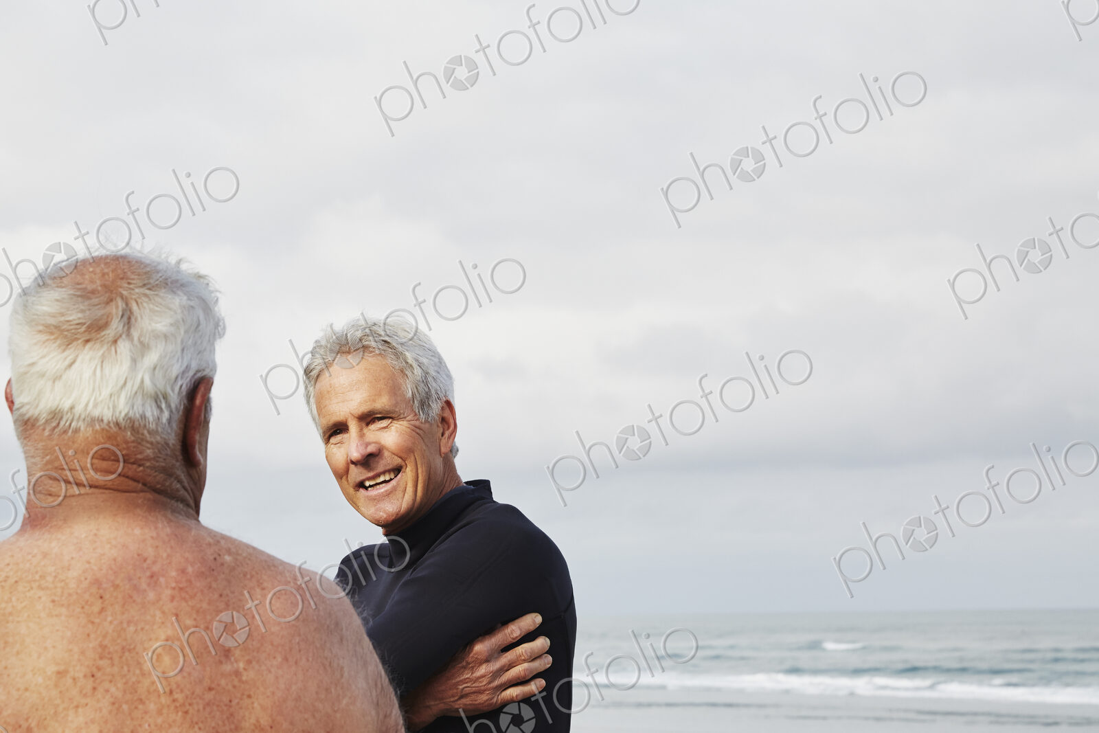 Two senior men standing on a beach chatting, one wearing a wetsuit.