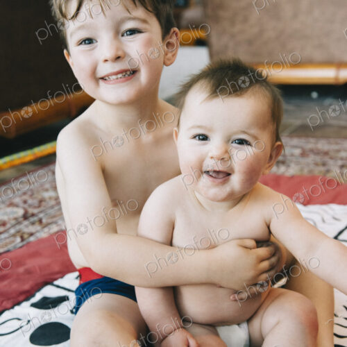 Two siblings, a boy and his baby sister sitting on a rug laughing.