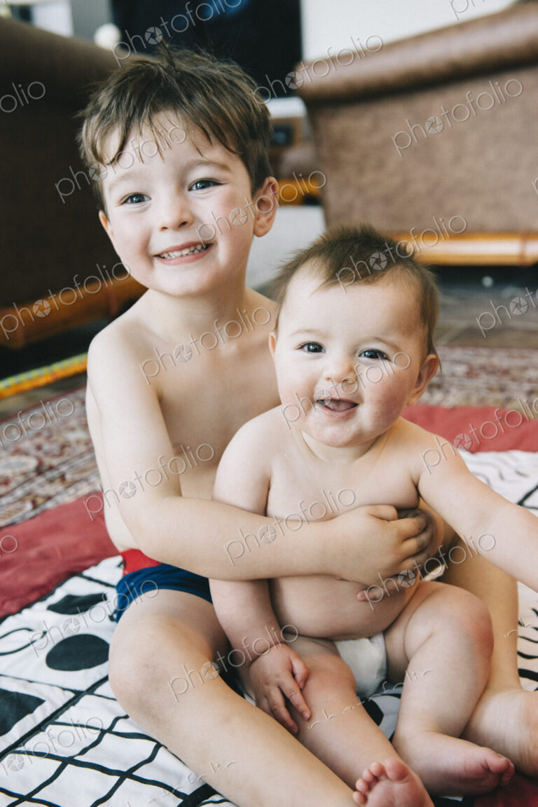 Two siblings, a boy and his baby sister sitting on a rug laughing.