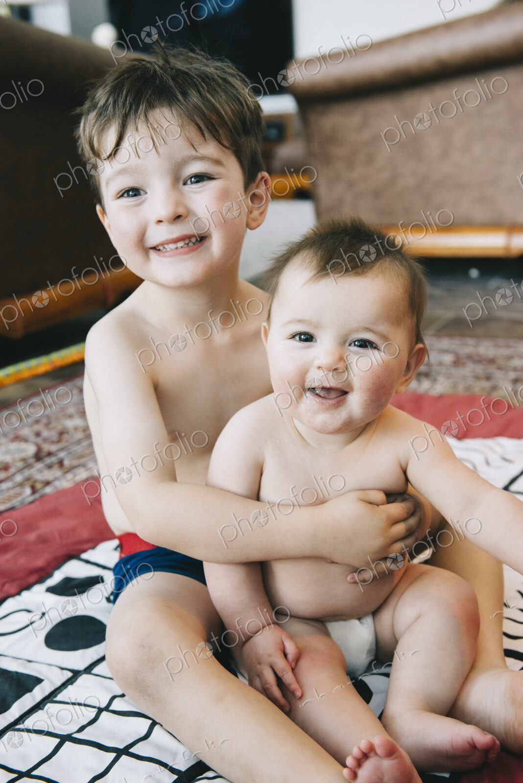 Two siblings, a boy and his baby sister sitting on a rug laughing.