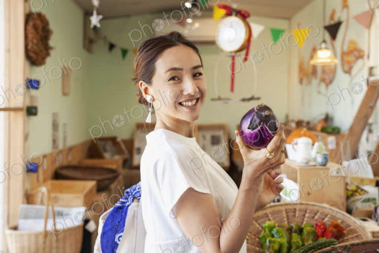 Two smiling Japanese women standing in a farm shop, holding aubergine, looking at camera.