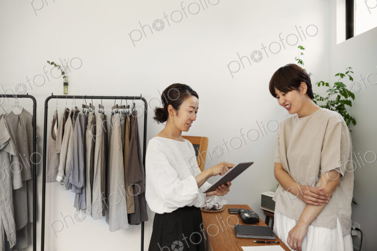Two smiling Japanese women standing in a small fashion boutique, holding digital tablet.