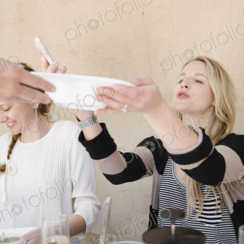 Two smiling women sitting at a table, one reaching for a plate of food.