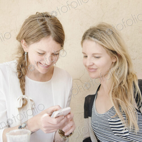 Two smiling women with long blond hair sitting at a table, looking at a cell phone.