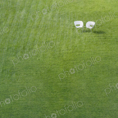 Two white office chairs on green lawn with pattern of stripes mown into grass meeting…