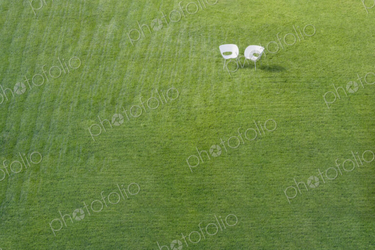 Two white office chairs on green lawn with pattern of stripes mown into grass meeting…