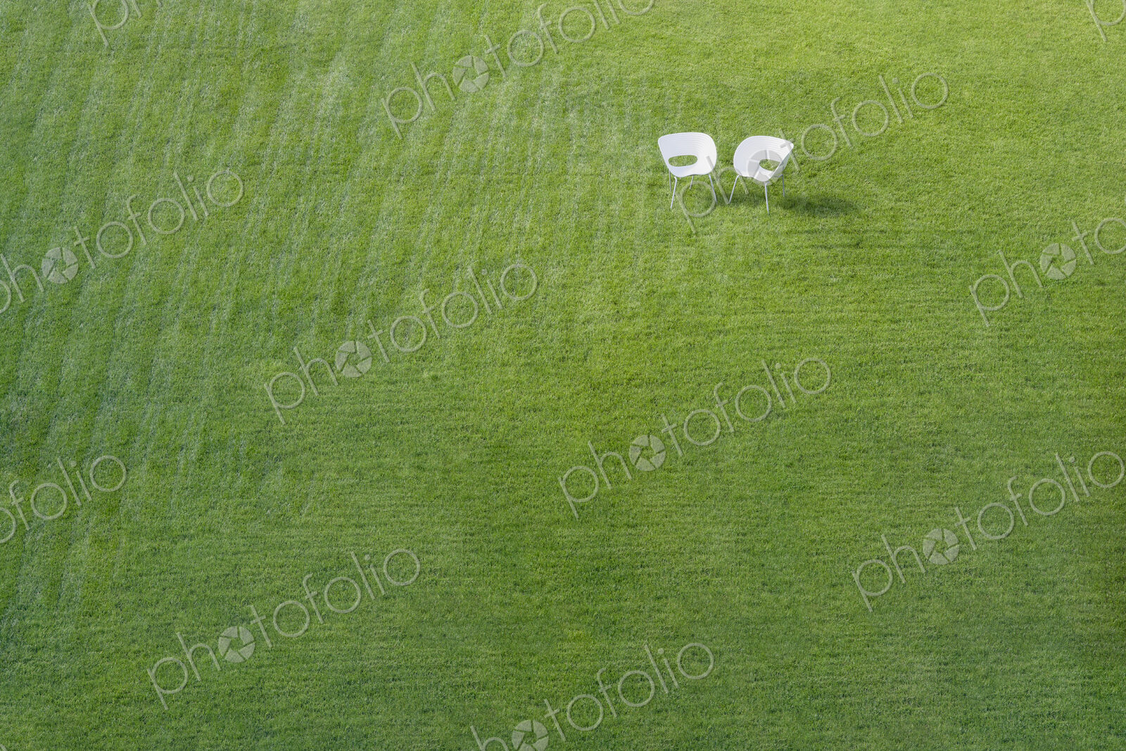 Two white office chairs on green lawn with pattern of stripes mown into grass meeting…