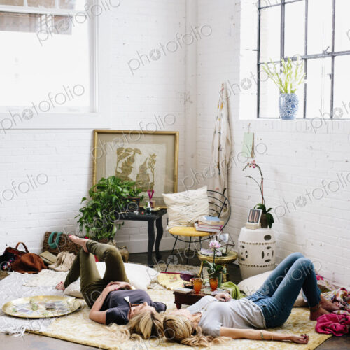 Two women lying on the floor of a room strewn with cushions and personal possessions. Two women lying on the floor of a room strewn with cushions and personal possessions.