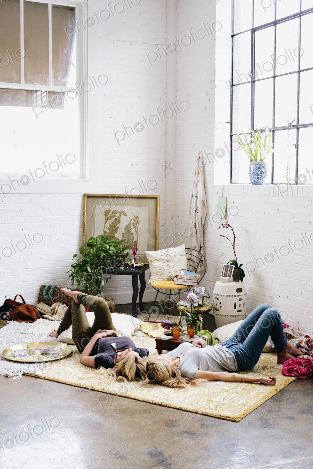 Two women lying on the floor of a room strewn with cushions and personal possessions.