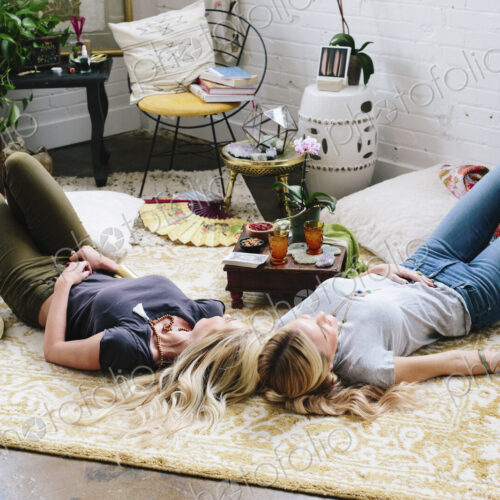 Two women lying on the floor of a room strewn with cushions and personal possessions. Two women lying on the floor of a room strewn with cushions and personal possessions.