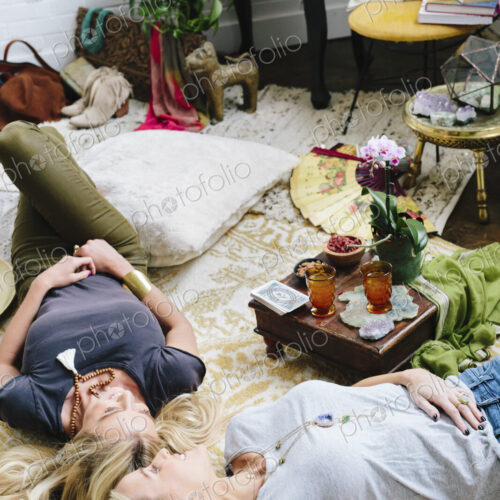Two women lying on the floor of a room strewn with cushions and personal possessions.