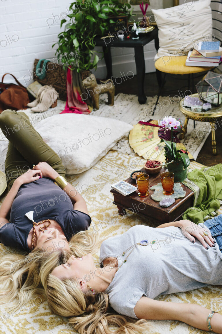 Two women lying on the floor of a room strewn with cushions and personal possessions.