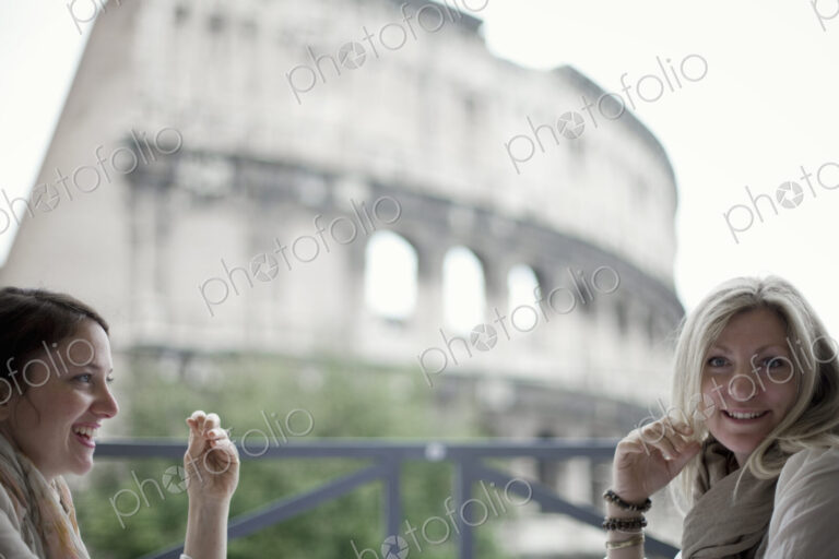 Two women seated on a terrace overlooking the huge Roman amphitheatre building, the…