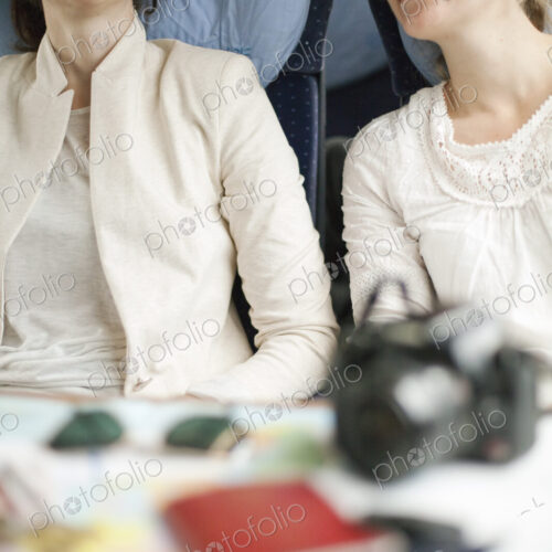 Two women seated on a train, side by side. Camera, passports and sunglasses on the…
