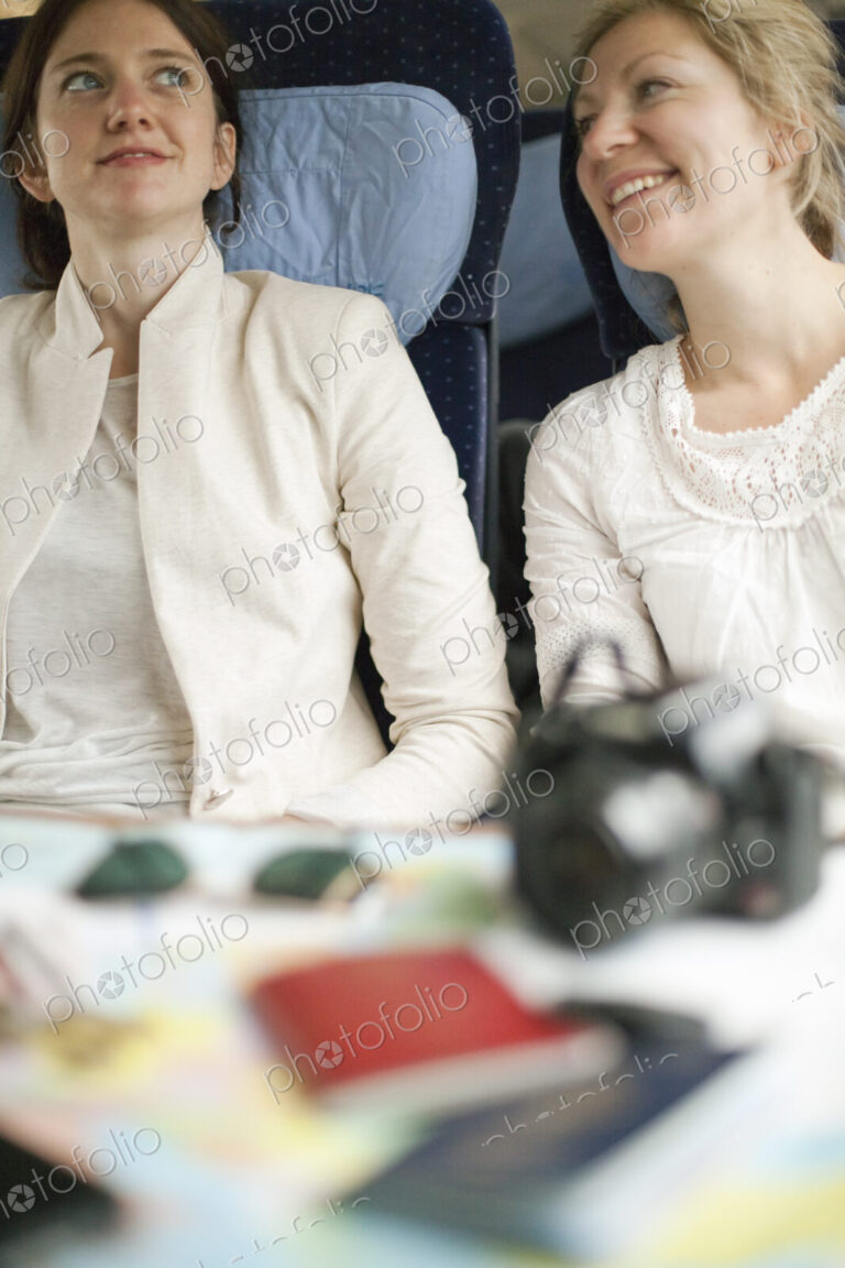 Two women seated on a train, side by side. Camera, passports and sunglasses on the…