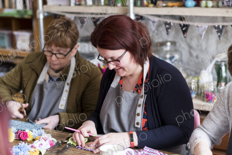 Two women sitting at a table in a workshop, making fabric flowers.