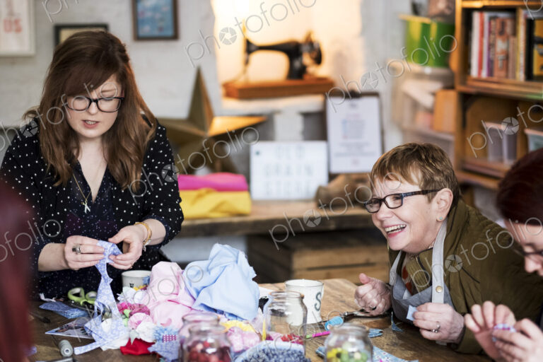 Two women sitting at a table in a workshop, making fabric flowers.