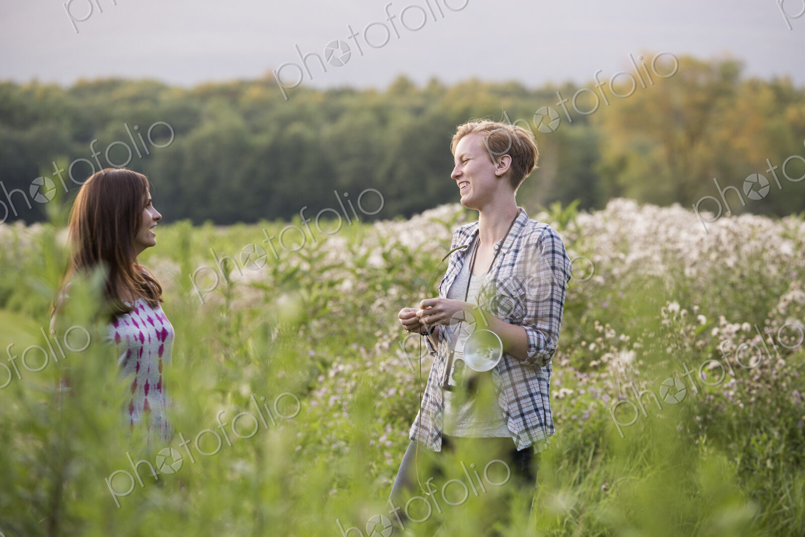 Two women standing in a meadow surrounded by tall grass and wild flowers.