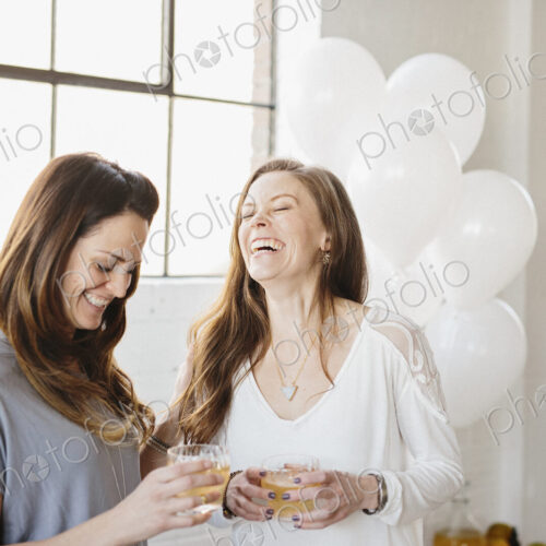 Two women standing side by side holding drinks. White balloons, party decorations. Two women standing side by side holding drinks. White balloons, party decorations.