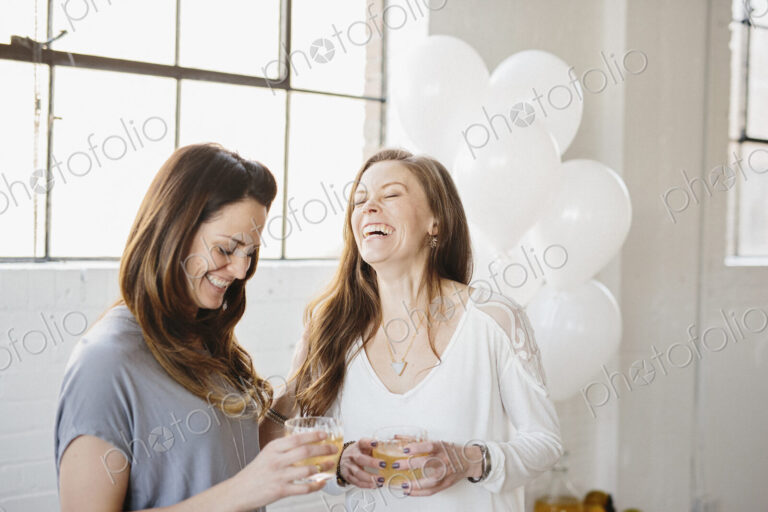 Two women standing side by side holding drinks. White balloons, party decorations.