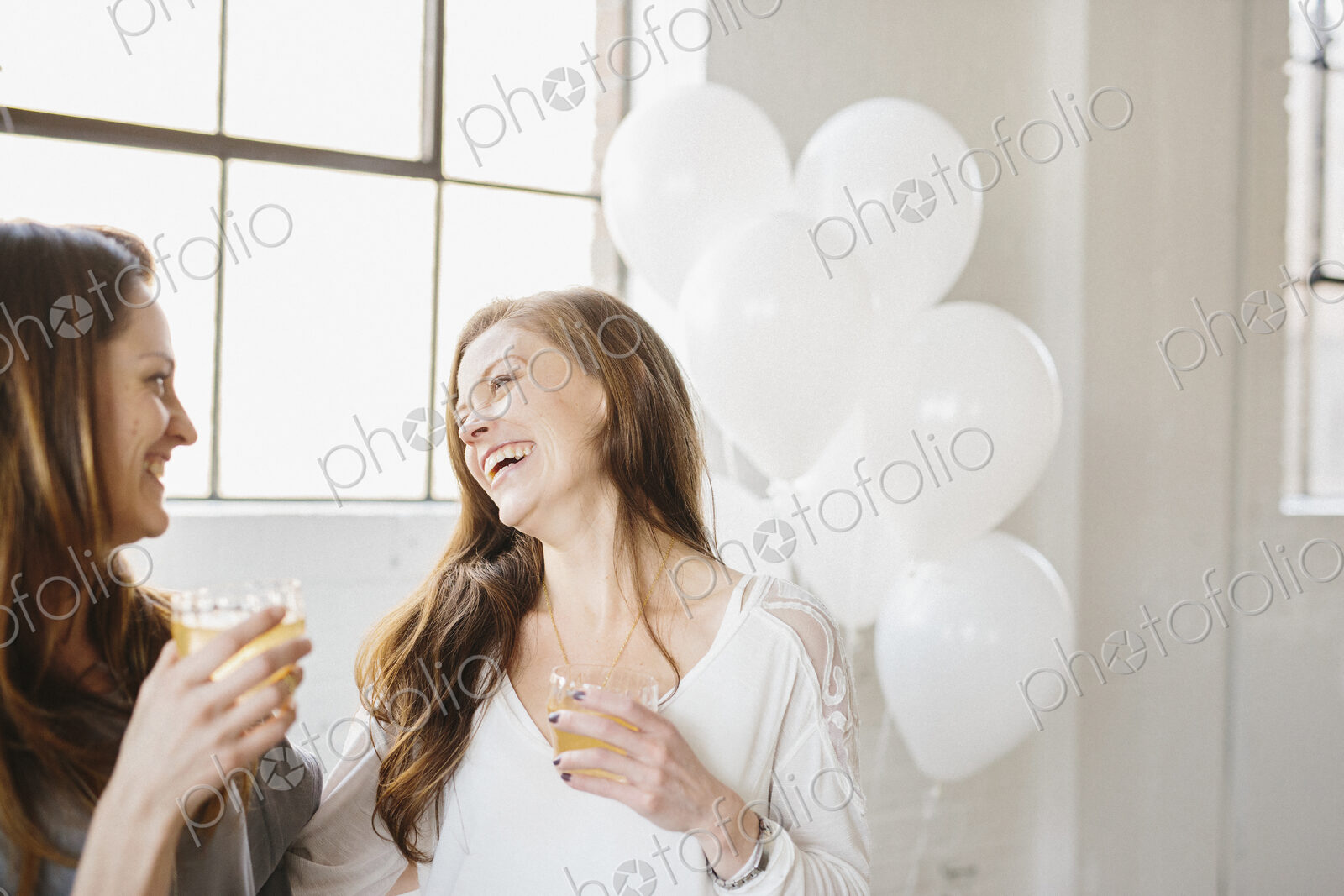 Two women standing side by side holding drinks. White balloons, party decorations.