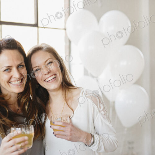 Two women standing side by side holding drinks. White balloons, party decorations. Two women standing side by side holding drinks. White balloons, party decorations.