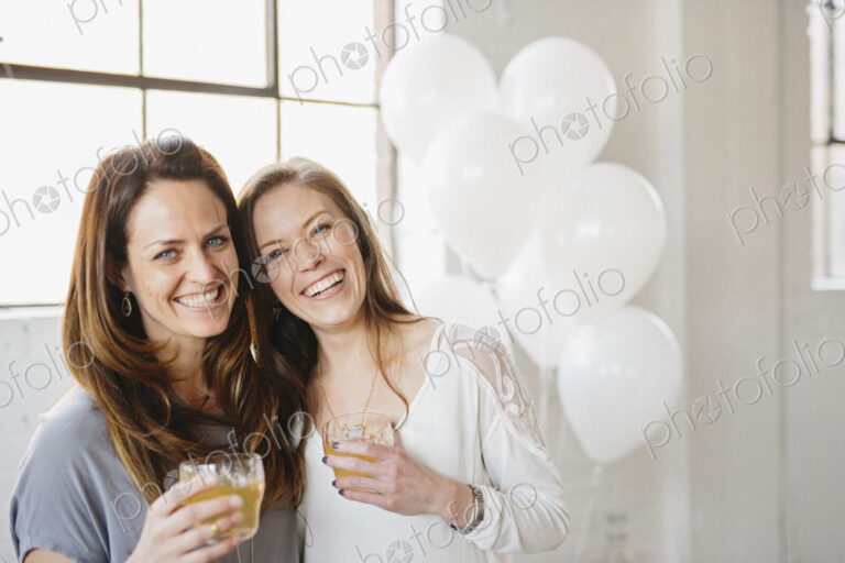 Two women standing side by side holding drinks. White balloons, party decorations.
