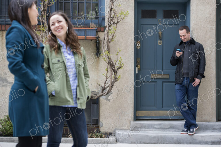 Two women talking as they walk down a street. A man leaning against a house door…