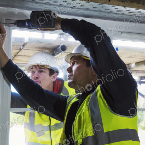 Two workmen on a construction site, builder in hard hat using an electric drill on a…