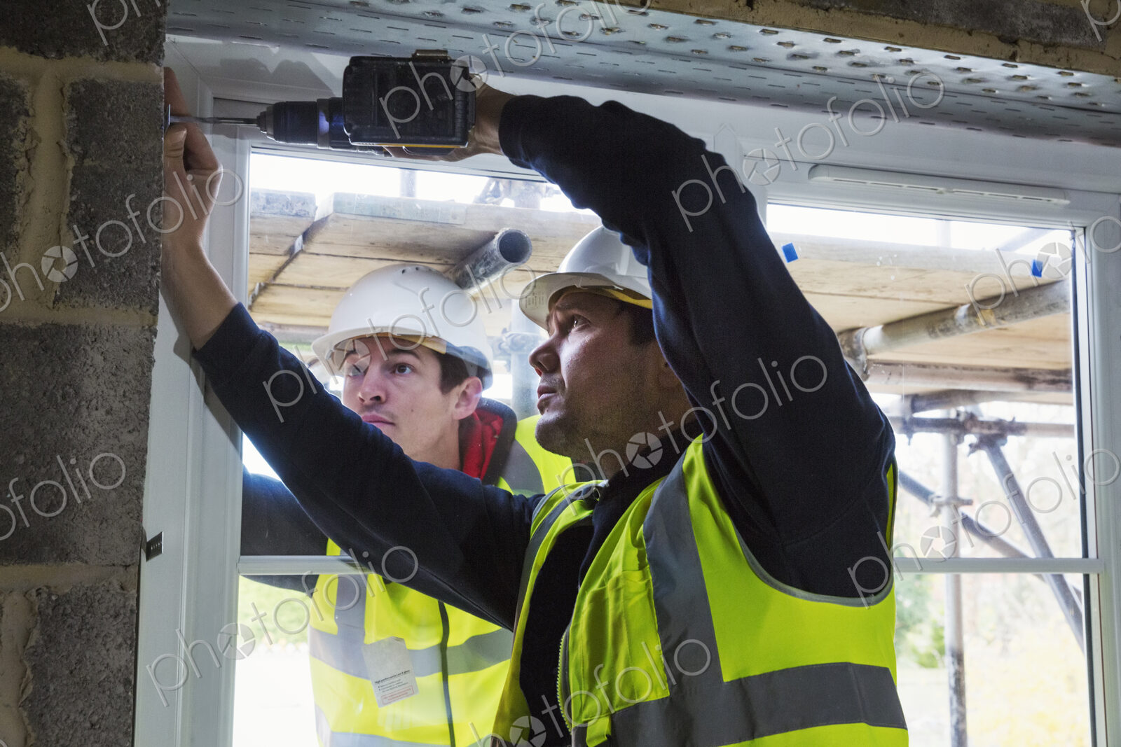 Two workmen on a construction site, builder in hard hat using an electric drill on a…