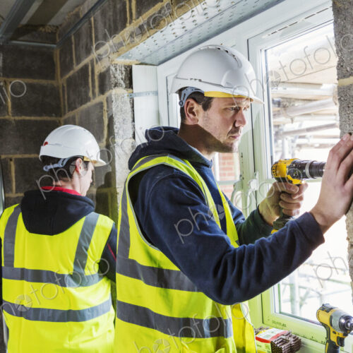 Two workmen on a construction site, builder in hard hat using an electric drill on a…
