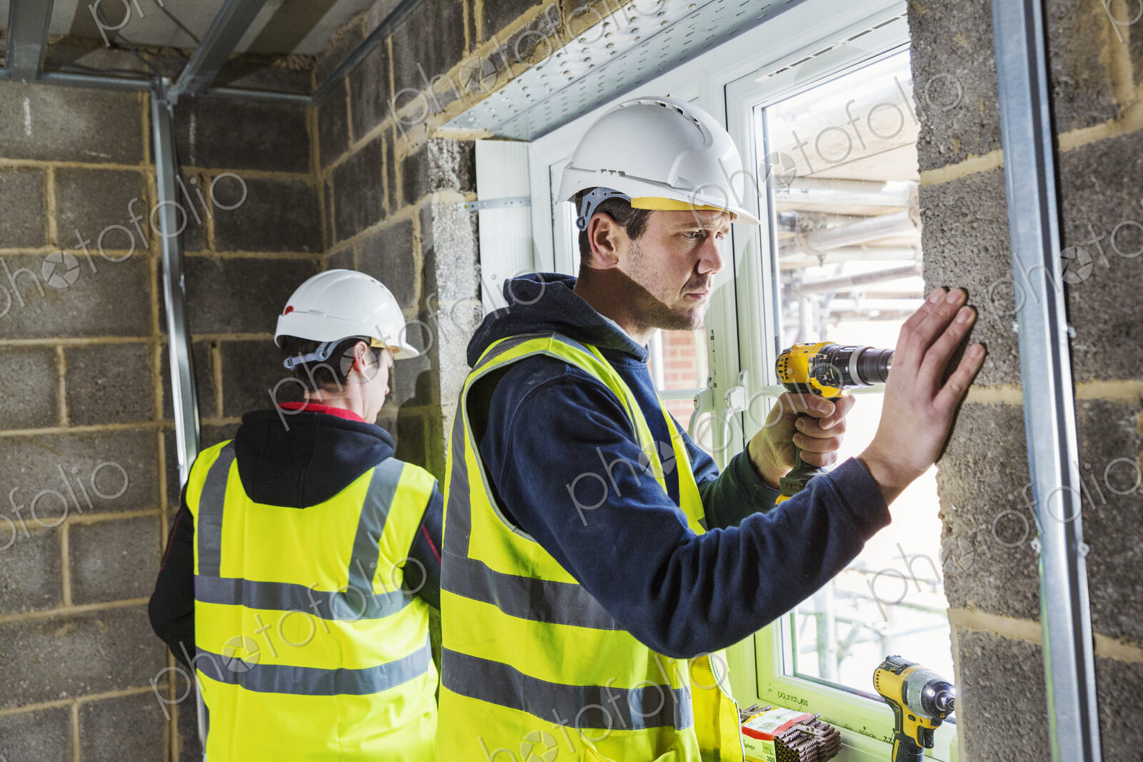 Two workmen on a construction site, builder in hard hat using an electric drill on a…
