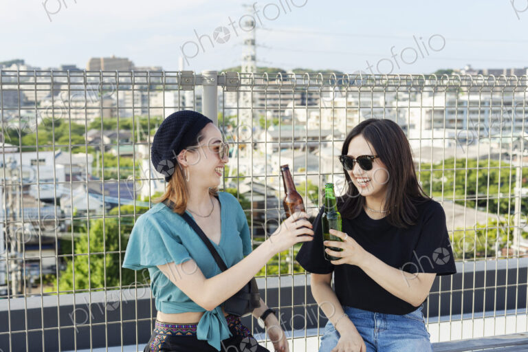 Two young Japanese women sitting on a rooftop in an urban setting, drinking beer.