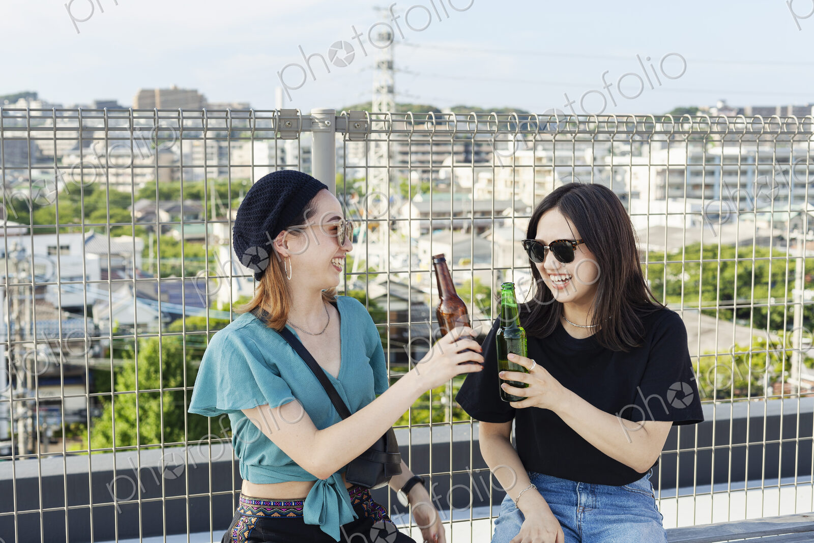 Two young Japanese women sitting on a rooftop in an urban setting, drinking beer.