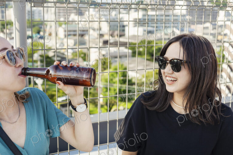 Two young Japanese women sitting on a rooftop in an urban setting, drinking beer.