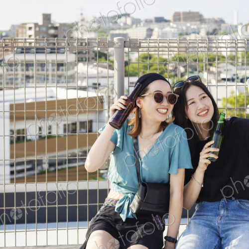 Two young Japanese women sitting on a rooftop in an urban setting, taking selfie with… Two young Japanese women sitting on a rooftop in an urban setting, taking selfie with…