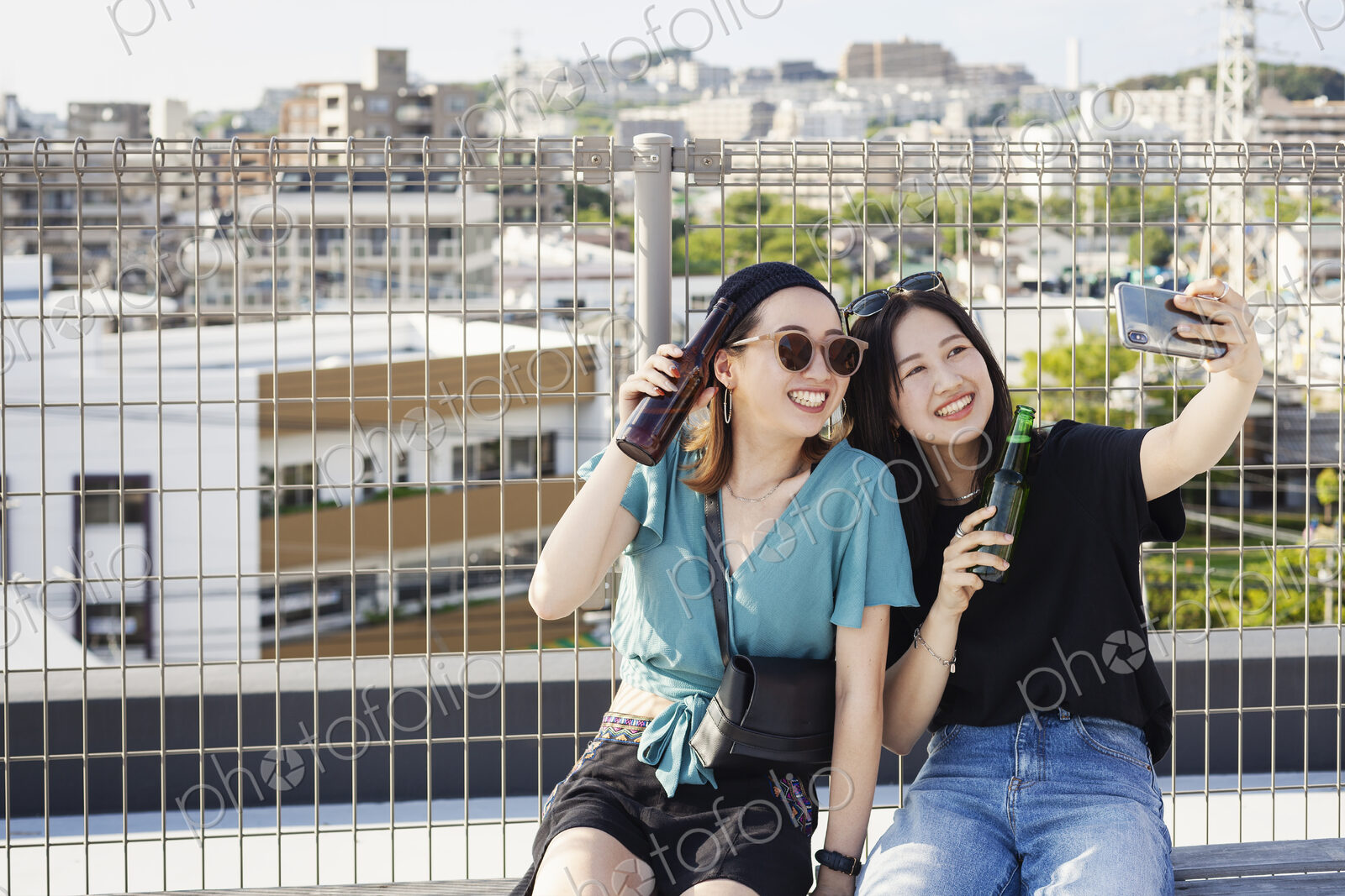 Two young Japanese women sitting on a rooftop in an urban setting, taking selfie with…