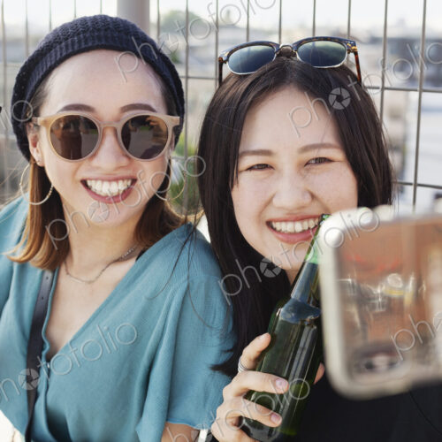 Two young Japanese women sitting on a rooftop in an urban setting, taking selfie with… Two young Japanese women sitting on a rooftop in an urban setting, taking selfie with…