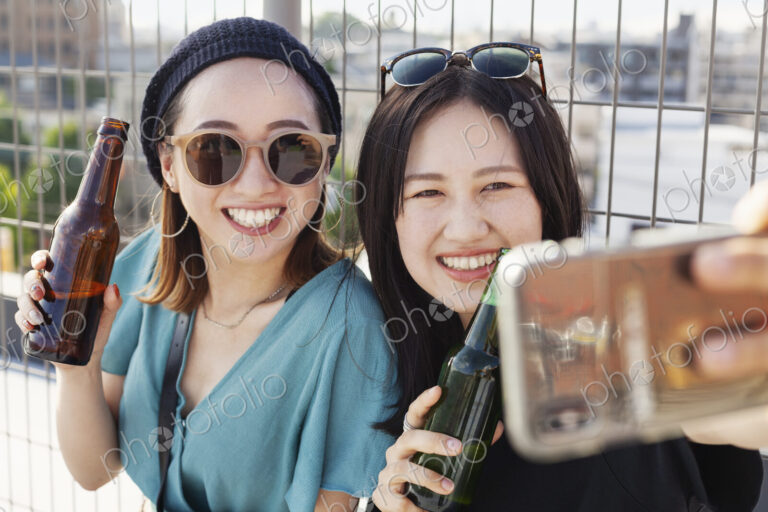 Two young Japanese women sitting on a rooftop in an urban setting, taking selfie with…