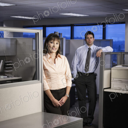 Two young business people working into the evening in a cubicle office set up. Two young business people working into the evening in a cubicle office set up.