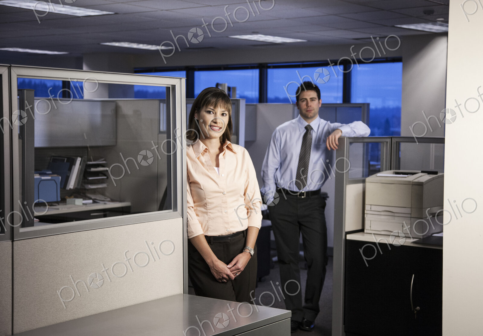 Two young business people working into the evening in a cubicle office set up.