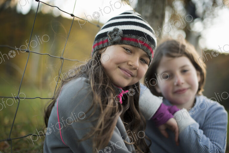 Two young girls side by side outdoors on a farm. Leaning on a fence post.