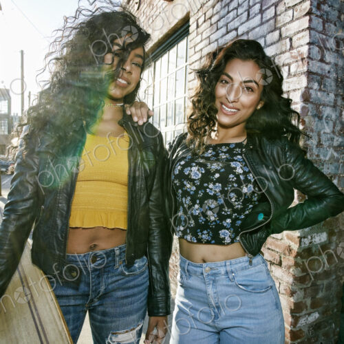 Two young smiling women with long curly black hair standing on pavement, holding…