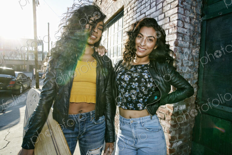 Two young smiling women with long curly black hair standing on pavement, holding…