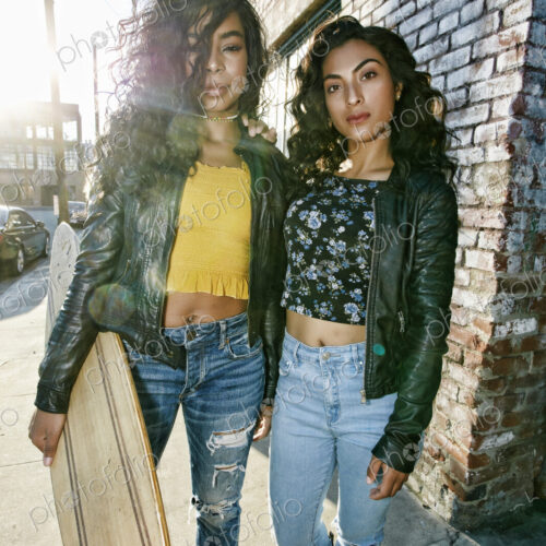 Two young smiling women with long curly black hair standing on pavement, holding…