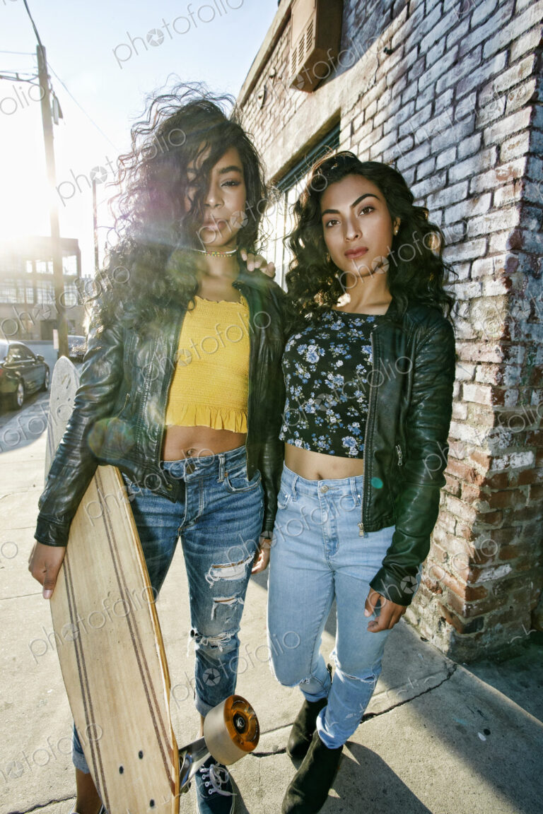 Two young smiling women with long curly black hair standing on pavement, holding…