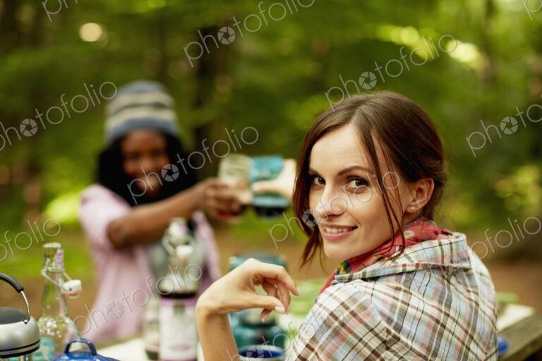 Two young woman drinking a toast at a picnic site or campsite in the woods.