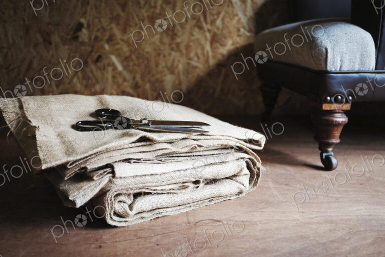 Upholstery workshop. Neutral coloured folded furnishing fabric, large scissors and a…