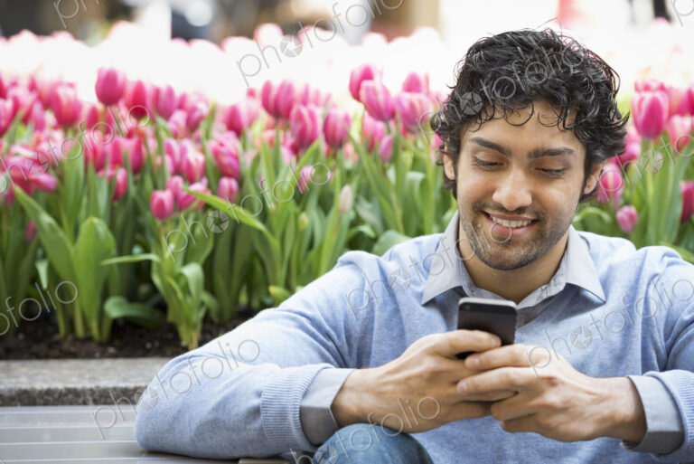 Urban Lifestyle. A man in the park, using his mobile phone. A bed of pink flowering…