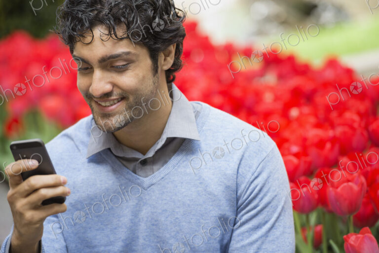 Urban Lifestyle. A man in the park, using his mobile phone. A bed of red flowering…